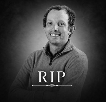 The image features a black and white photo of a man with curly hair wearing a gray sweater, smiling for the camera. The caption below the photo reads "RIP" which is written in white text. The background of the photo appears to be a dark color, possibly a wall or a piece of furniture.
