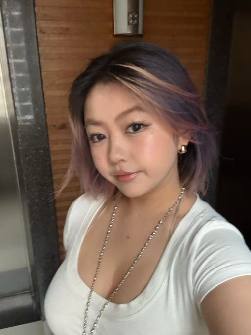 A selfie of a young woman with purple-tinted hair wearing a white top and a chain necklace.