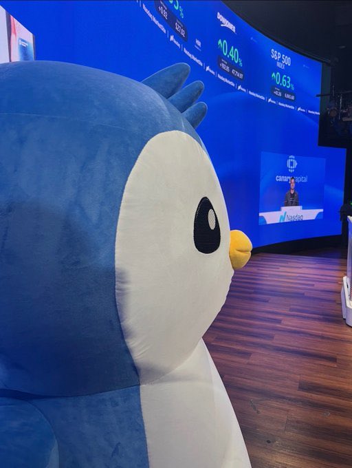A giant plush Pudgy Penguin sits prominently in the foreground at what appears to be a Nasdaq event at the Canyon Capital building, with a curved digital screen in the background displaying "S&P 500" and "+0.40%" market data alongside stock indices like "60.85" and "9,720.99," humorously suggesting that owning one of these crypto-native mascots might be the secret pass into the elite world of high finance.