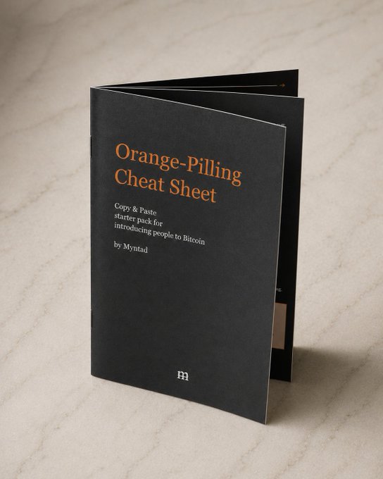 A black booklet titled 'Orange-Pilling Cheat Sheet - Copy & paste starter pack for introducing people to Bitcoin by Myntad' displayed standing up on a marble surface.