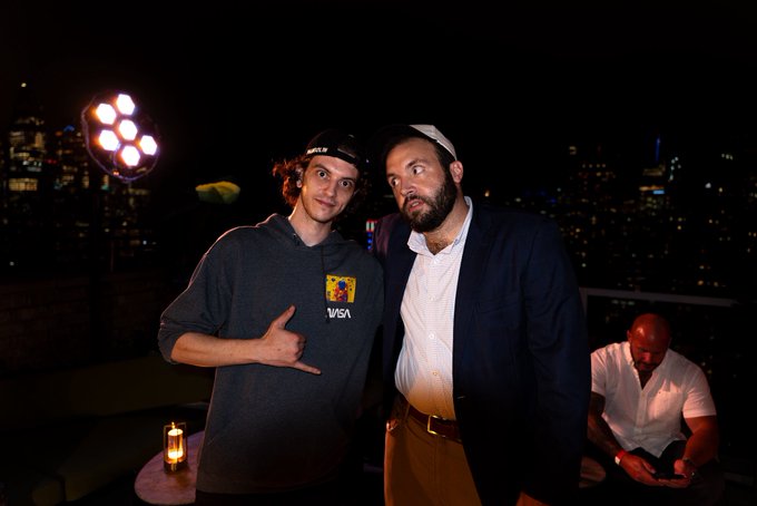 Two men pose for a photo at a nighttime rooftop party with a city skyline behind them—one in a gray hoodie giving a 'shaka' sign, the other in a blue blazer with a beard and headband, while a third person sits in the background.