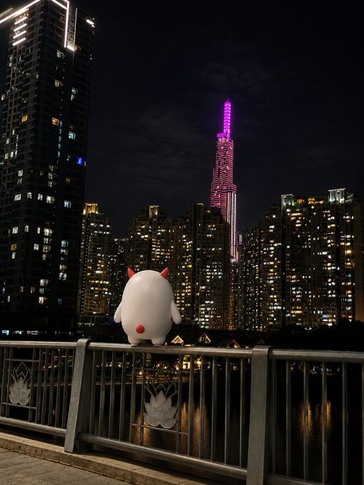 A nighttime cityscape featuring a small white plush character with red horns perched on a railing, overlooking a river with a tall illuminated skyscraper glowing pink in the background.