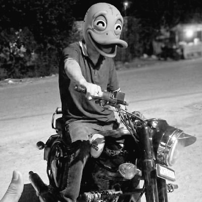 The image features a person wearing a duck mask while sitting on a motorcycle. The person appears to be smiling, possibly posing for a photo or enjoying their time on the bike. The scene takes place at night, adding an element of mystery and intrigue to the overall composition.