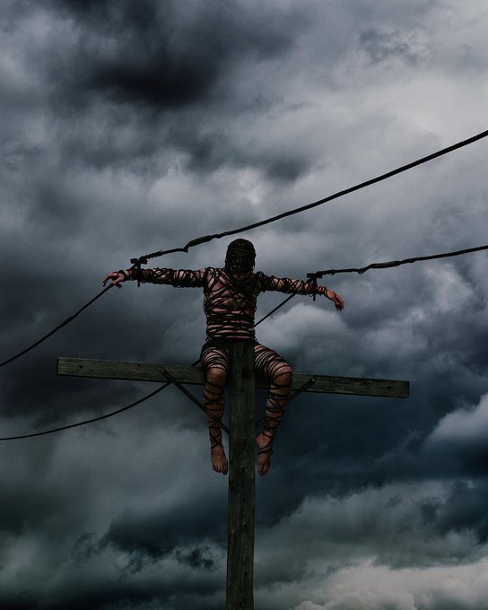 A dramatic photograph of a figure tied to a wooden cross with ropes against a dark, stormy cloudy sky, with cables stretching across the scene.