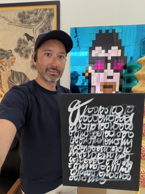 A man wearing a navy t-shirt and a cap takes a selfie while holding a black canvas with white calligraphic-style lettering, standing in front of two other artworks including a pixelated portrait.