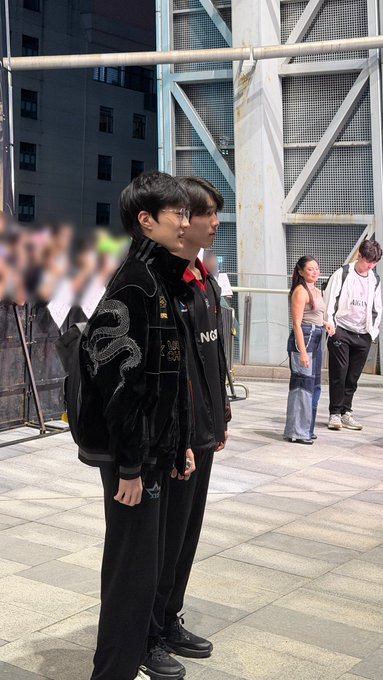 Two young men in black outfits stand together outdoors on a paved area, one wearing a jacket featuring a dragon motif.