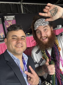 Two men posing together at a press event, one wearing a bandana and colorful attire with a hand gesture behind his head, alongside a 'PRESS MEDIA' banner.