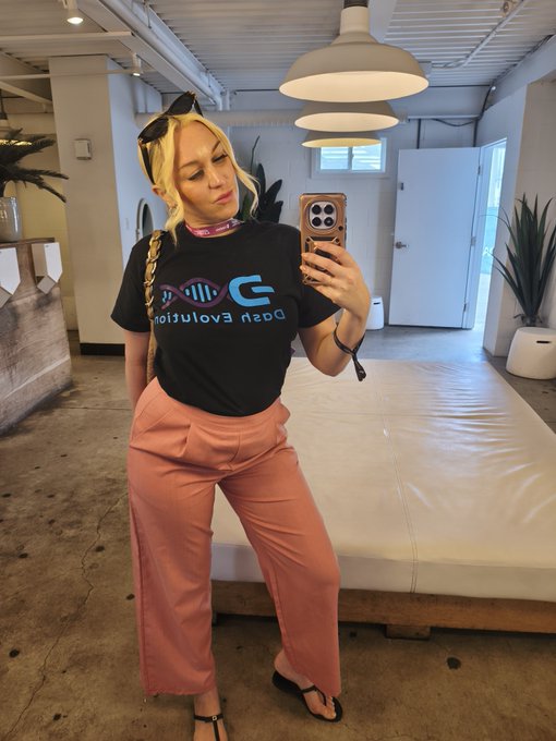 Mirror selfie of a blonde woman with braided hair wearing sunglasses on her head, a black 'open evolution' t-shirt, and pink pants in an industrial-style room.