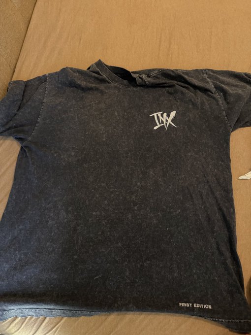 A faded black/gray acid-wash t-shirt with a stylized 'JB' logo on the chest and 'FIRST EDITION' text near the bottom hem, laid flat on a brown surface.