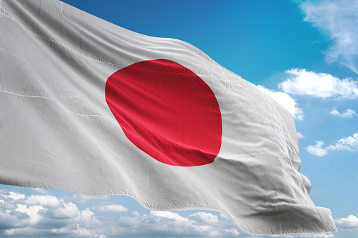 Japan’s flag waves against a blue sky, symbolizing the country as it explores blockchain-based settlement for stocks and government bonds, with a formal plan expected by 2027.