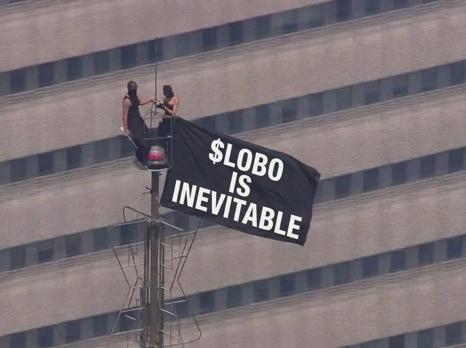 Two people on an elevated platform against a skyscraper displaying a black banner that reads '$LOBO IS INEVITABLE'.