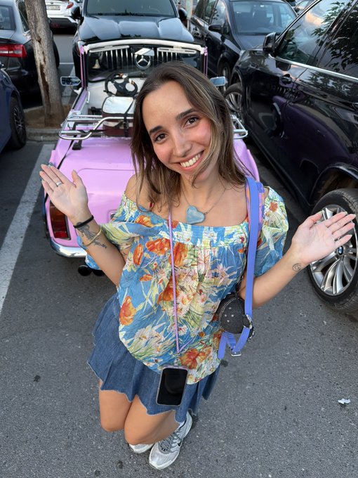 A smiling woman in a floral off-shoulder top and denim skirt poses in front of a pink open-top Mercedes-Benz car parked on a street.