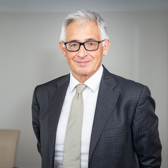 A professional headshot of a smiling older man with gray hair and glasses, wearing a gray plaid suit, white shirt, and patterned tie.
