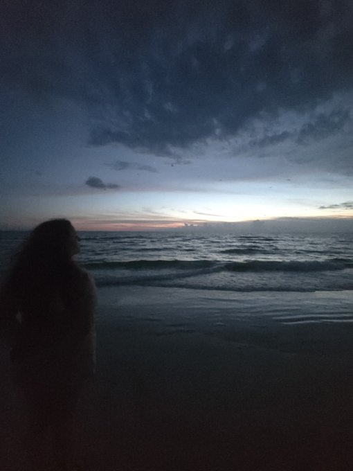 A solitary woman stands on a beach silhouetted against a twilight ocean, gazing out at the waves as the sun sets behind the horizon. The serene, contemplative scene contrasts with the post's message about valuing a genuine, faith-driven partner who won't be swayed by the promise of cryptocurrency wealth.