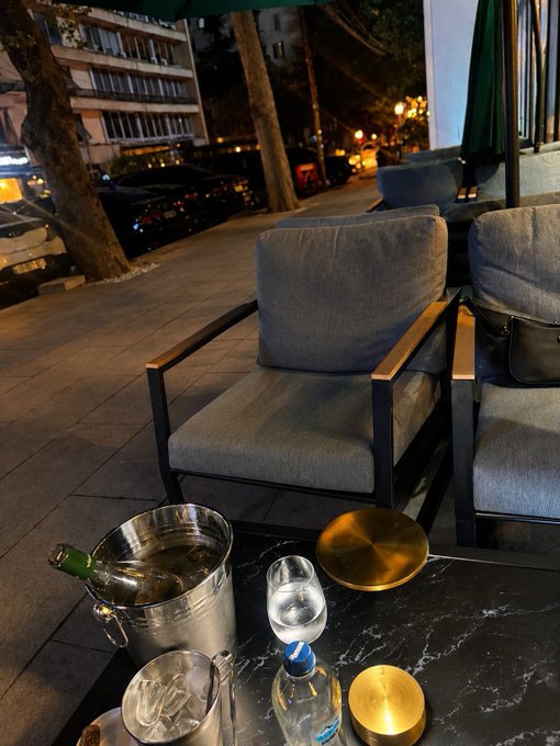 A nighttime view from an outdoor patio shows a black marble table with a wine bucket, water bottles, and glasses, with gray armchairs and parked cars visible on the street below.