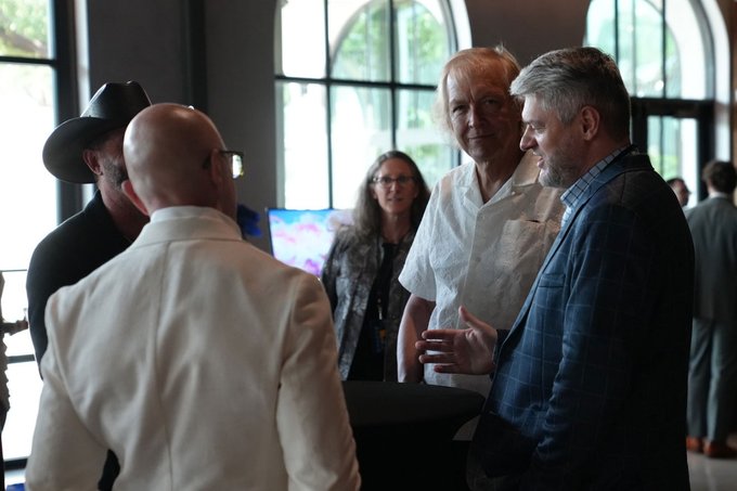 A photograph of a group of people conversing at what appears to be an indoor social event or conference, featuring a man in a cowboy hat, a bald man in a white suit, a woman in a white blouse, and a man in a blue checkered blazer.