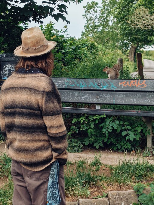 A person in a frayed straw hat and striped sweater stands before a graffiti-covered bench where a squirrel perches, surrounded by lush greenery—while visible text on the bench reads "SUCH THINGS VENOMS ZILT THV" alongside tags like "Hank" and other scrawled words, echoing the post's reflective mood about impermanence and loss.