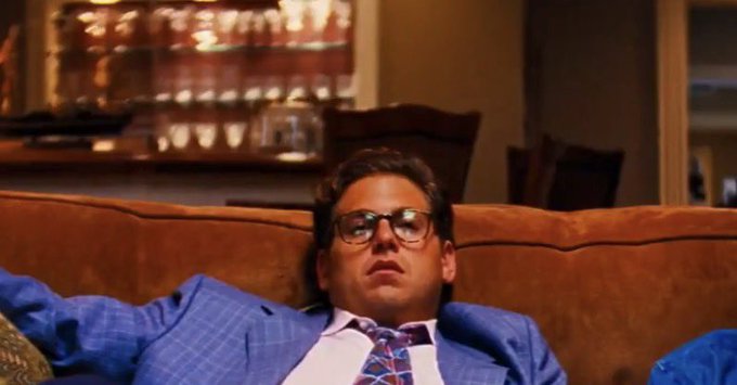 A man in glasses, a blue suit, and a patterned tie slumps exhausted on a brown couch, conveying the weary frustration implied by the post's critique of Coinbase halting their monthly ICOs after Monad and warning employees not to transfer or sell the tokens.