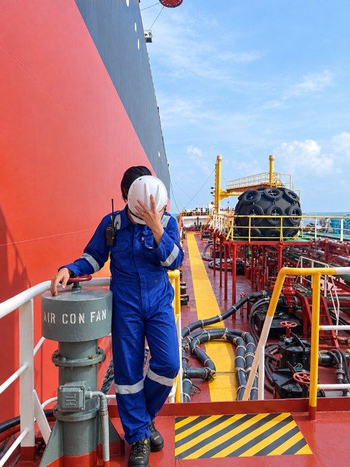 A worker in a blue coverall and white hard hat stands on the deck of a large red and blue ship, holding onto a ventilation structure labeled 'AIR CON FAN', with industrial equipment and ocean in the background.