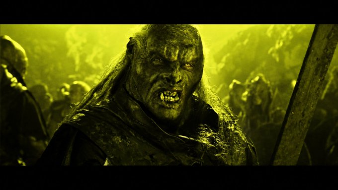 A cinematic still of a menacing, pale-skinned creature with long hair, fangs, and glowing eyes in a yellowish-green environment, likely a fantasy or horror film scene.