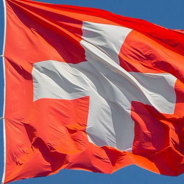 A close-up photograph of a red flag with a white cross, the national flag of Switzerland, waving against a clear blue sky.