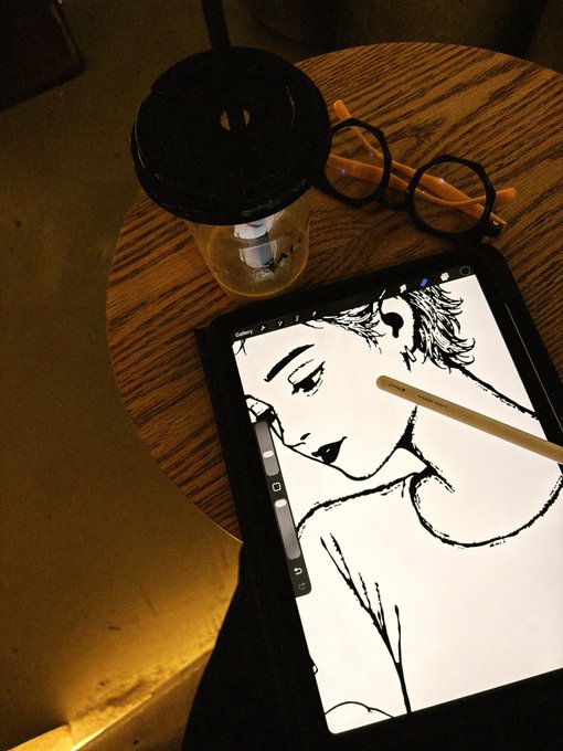 A tablet displaying a black and white digital illustration of a person, accompanied by a stylus, placed on a wooden table alongside a plastic cup with a lid and a pair of orange-rimmed glasses.