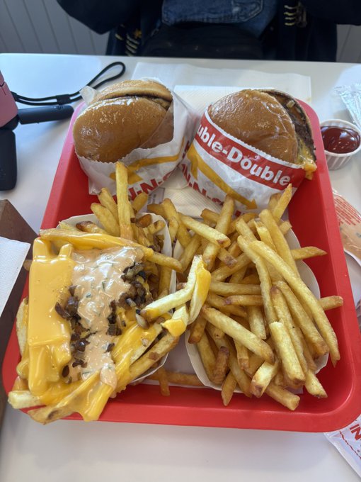 A red tray on a table holds two burgers in paper wrappers reading 'Double', a pile of french fries, and a generous serving of fries covered in melted cheese sauce.