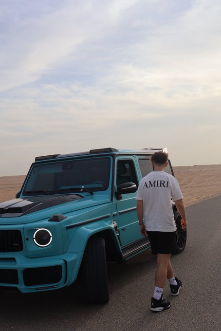 A man wearing an "AMIRI" t-shirt walks toward a turquoise Mercedes G-Wagon parked on a desert road, matching the post's casual Sunday greeting.