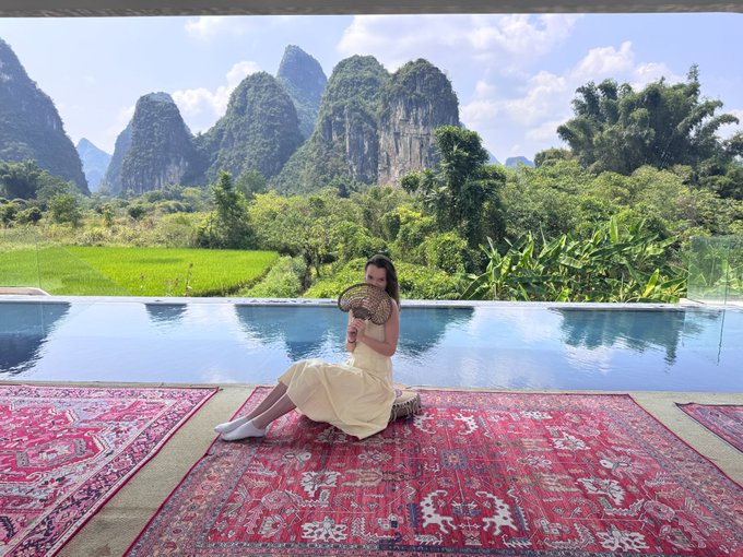 A woman sits on ornate red rugs by an infinity pool with a stunning view of karst mountains and lush green rice fields in the background, fanning herself.