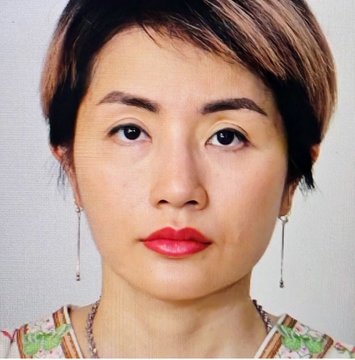 A portrait photograph of a woman with short dark hair wearing dangling earrings and a floral patterned top, with red lipstick and a neutral expression against a light background.
