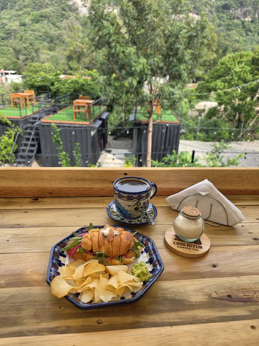 A meal of croissants and chips served on a decorative plate with a cup of coffee on a wooden table at Churyuc Coffee, overlooking a green forested area with shipping container structures.