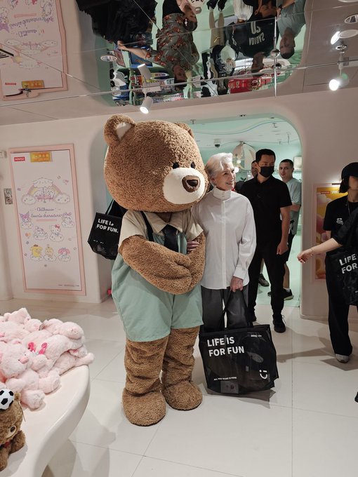 A large brown bear mascot wearing a teal outfit standing in a store next to a person with white hair, with other people in black masks and 'LIFE IS FOR FUN' bags visible.