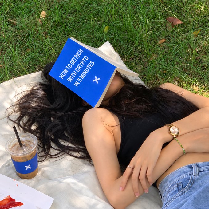 A woman lies on a blanket in the grass, using a blue book titled "HOW TO GET RICH WITH CRYPTO IN 5 MINUTES" as a pillow to shield her face from the sun, accompanied by an iced coffee cup—perfectly capturing the "gm, today we read first" vibe of leisure learning.
