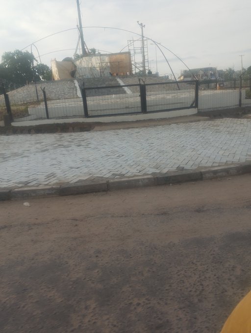 A street view showing a fenced compound with water tanks and an arched structure under construction behind a tiled walkway and dirt road.