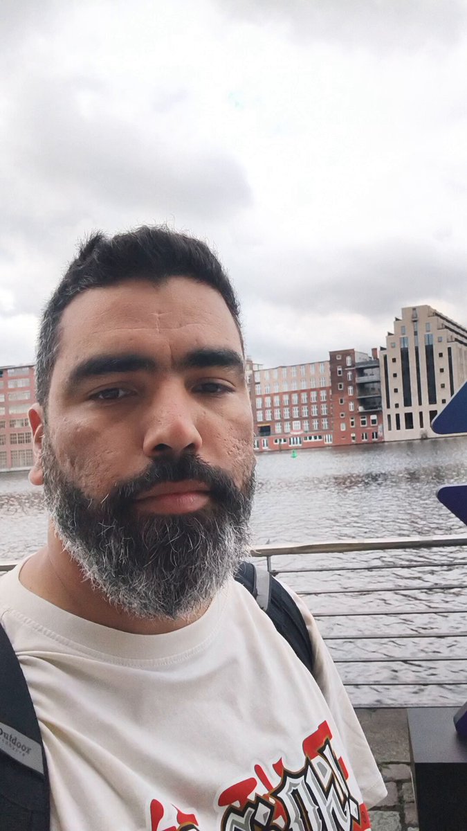 The image features a man with a beard wearing a white t-shirt, standing near a body of water or river. He has his head tilted back as if posing for a photo. The background includes buildings, suggesting that he might be at an urban location. There is also text in the image, which reads "Hey @solana summit is here LFG!!! @SuperteamDE". This implies that there could be a social media event or gathering happening nearby, possibly related to the #solana summit.