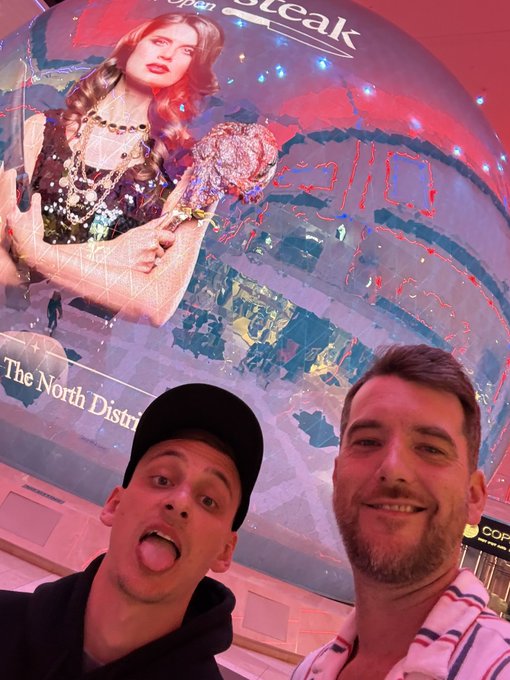 Two men taking a selfie in front of a large circular 'Golden Steer' steakhouse advertisement featuring a woman holding a large tomahawk steak, with 'The North District' visible below.