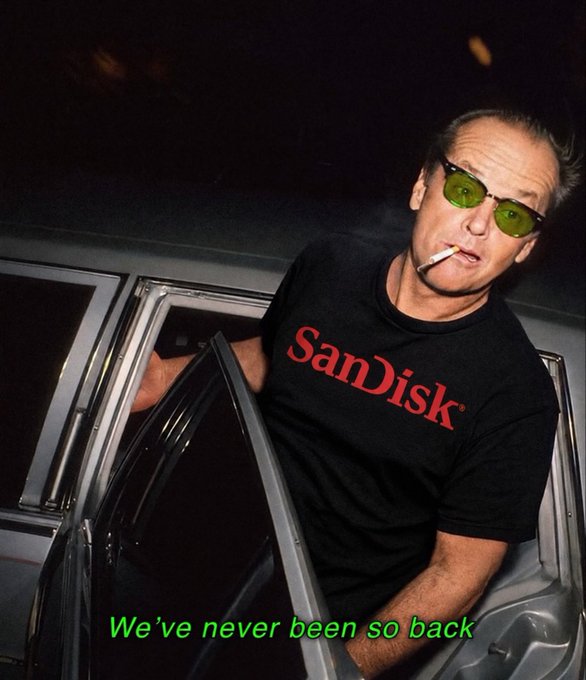 A stylized image of a man with green-tinted sunglasses smoking a cigarette while wearing a black 'SanDisk' t-shirt, exiting a car at night, with green text overlay reading 'We've never been so back.'