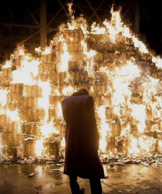 A lone figure in a long coat stands with their back turned before a massive, towering bonfire of stacked crates or pallets engulfed in flames, evoking destruction and loss. The image powerfully mirrors the post's sentiment of watching one's crypto investments burn after a devastating crash.