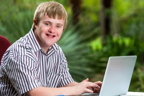A young man with short blonde hair, wearing a striped shirt, smiles at the camera while using a silver laptop in an outdoor setting with green foliage behind him.