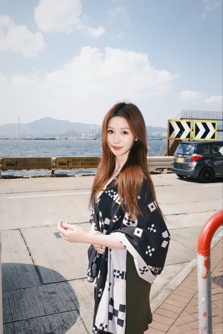 A young woman with long brown hair stands on a waterfront promenade, wearing a black and white patterned kimono-style top, with mountains and a harbor visible in the background.