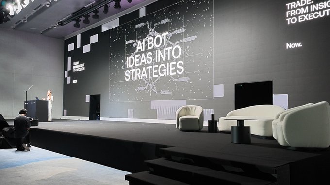 A large conference stage with a digital backdrop displaying 'AI BOT IDEAS INTO STRATEGIES', featuring a speaker at a podium and white lounge furniture on the right.