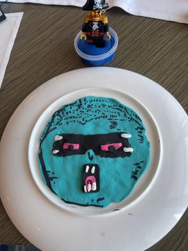 The image features a white plate with a blue face drawn on it, which appears to be made out of play dough or clay. The plate has a small toy pirate figurine sitting next to it. In the background, there are two cups - one is filled with a blue substance and the other contains a green substance.