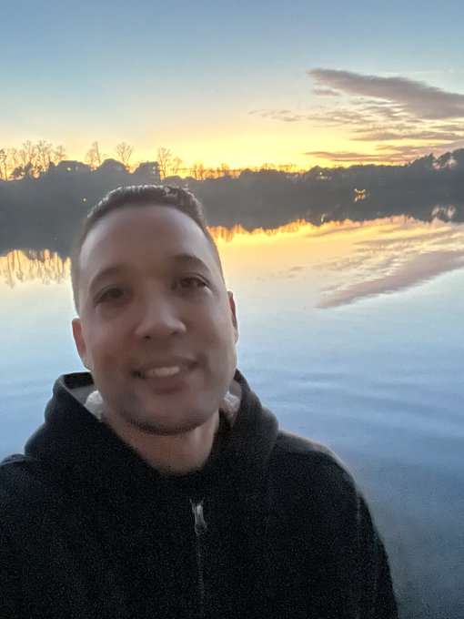 A selfie of a smiling man wearing a dark hooded sweatshirt, with a beautiful sunset reflecting over a calm lake behind him.