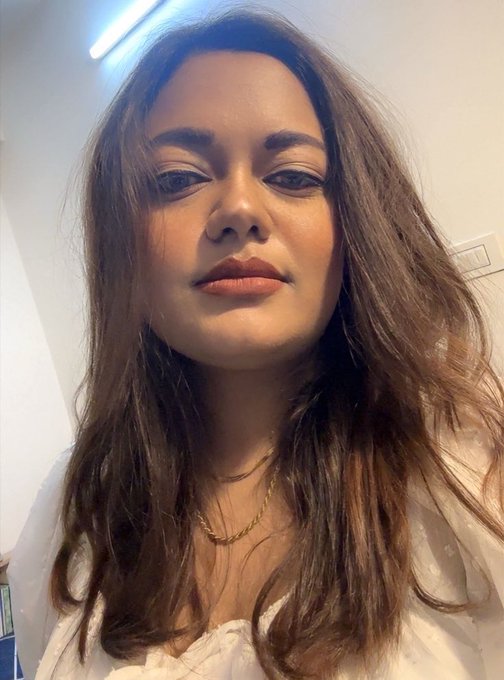 A selfie portrait of a young woman with long, wavy brown hair wearing a white top and a gold necklace, photographed indoors against a white wall.