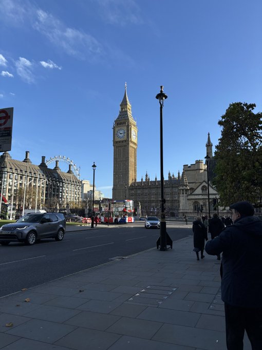 Street view of London's Big Ben and Westminster on a sunny day, with a London Eye visible in the background, a red double-decker bus, and pedestrians on the sidewalk.