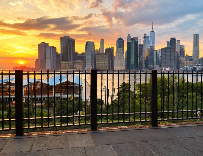 A scenic sunset view of a city skyline, likely Lower Manhattan, taken from across a body of water and framed by a black metal fence in the foreground.