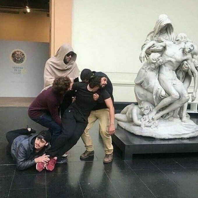 A group of young people, including one in a Ghostface-like hood, playfully pose together in front of a classical marble sculpture of multiple figures in a museum setting.