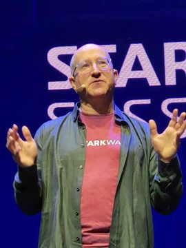 A bald man with glasses, wearing a green jacket over a pink "DARKWAVE" t-shirt, gestures with both hands while speaking on stage in front of a blue backdrop displaying partial text reading "STAR...". The image relates to the post about Zcash co-founder forecasting $ZEC reaching $5,000 by end of 2026.