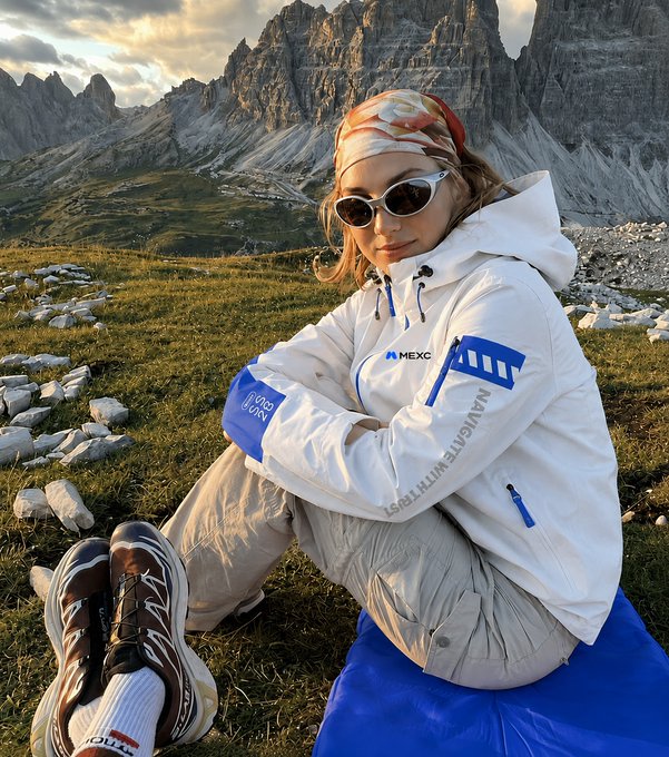 A woman in a white MEXC-branded puffer jacket, sunglasses, and a flame-patterned bandana sits on a blue cushion in an alpine meadow, posing with rugged Dolomites peaks towering behind her. The jacket prominently displays "MEXC" and the slogan "NAVIGATE WITH EASE," tying into the post's theme of chasing a new "ATH" (all-time high) through outdoor adventure and mountain scenery.