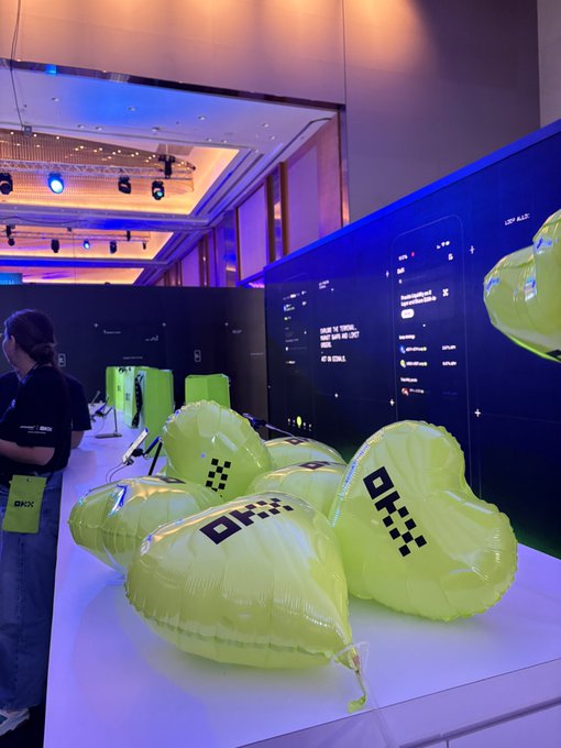 An event display showing several green heart-shaped balloons and boxes branded with the "OKX" logo, with a large screen in the background.